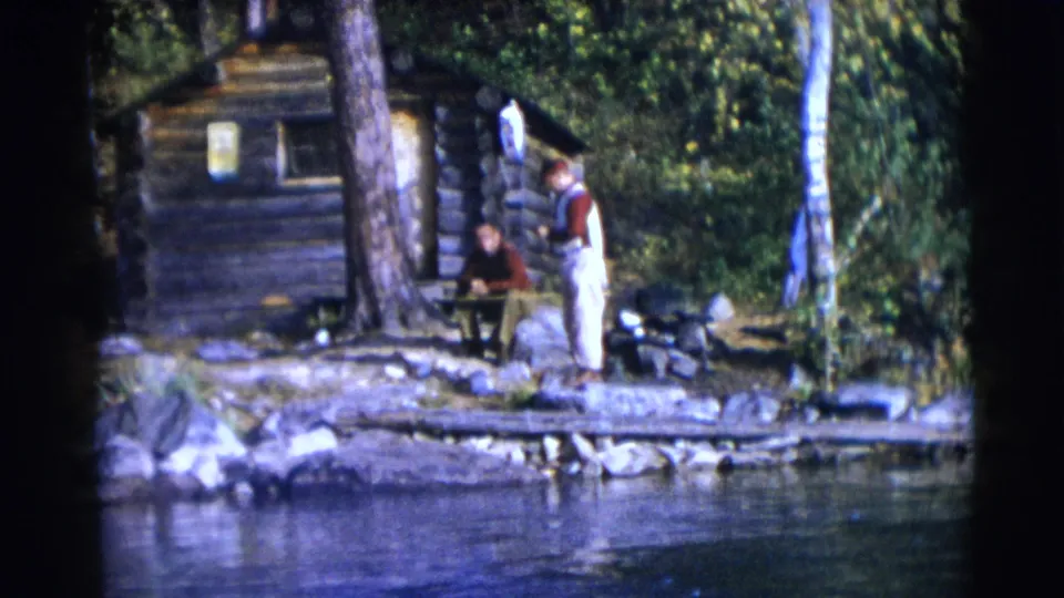 1953 Baraboo home-movie frame showing two men near a lakeside log cabin with trees, rocks, and water in the foreground.