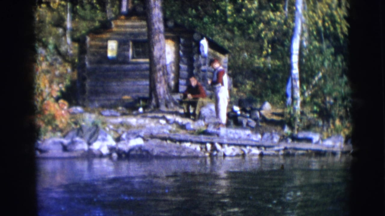 A man stands near a seated man outside a log cabin by the water.