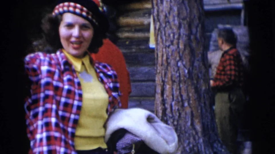 A smiling woman in a red, white, and blue tartan jacket and matching hat stands near a log cabin in the 1953 Baraboo footage.
