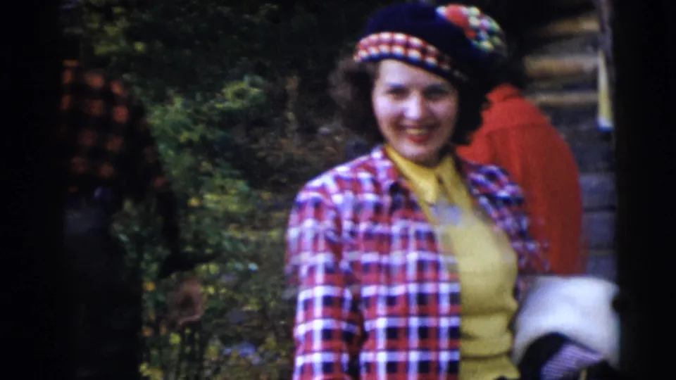 The woman in tartan clothing faces the camera near the Baraboo lakeside cabin, smiling with adults partly visible behind her.