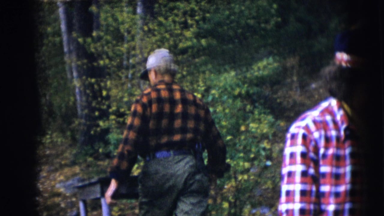Man in orange plaid flannel walks through woods as another person follows nearby.