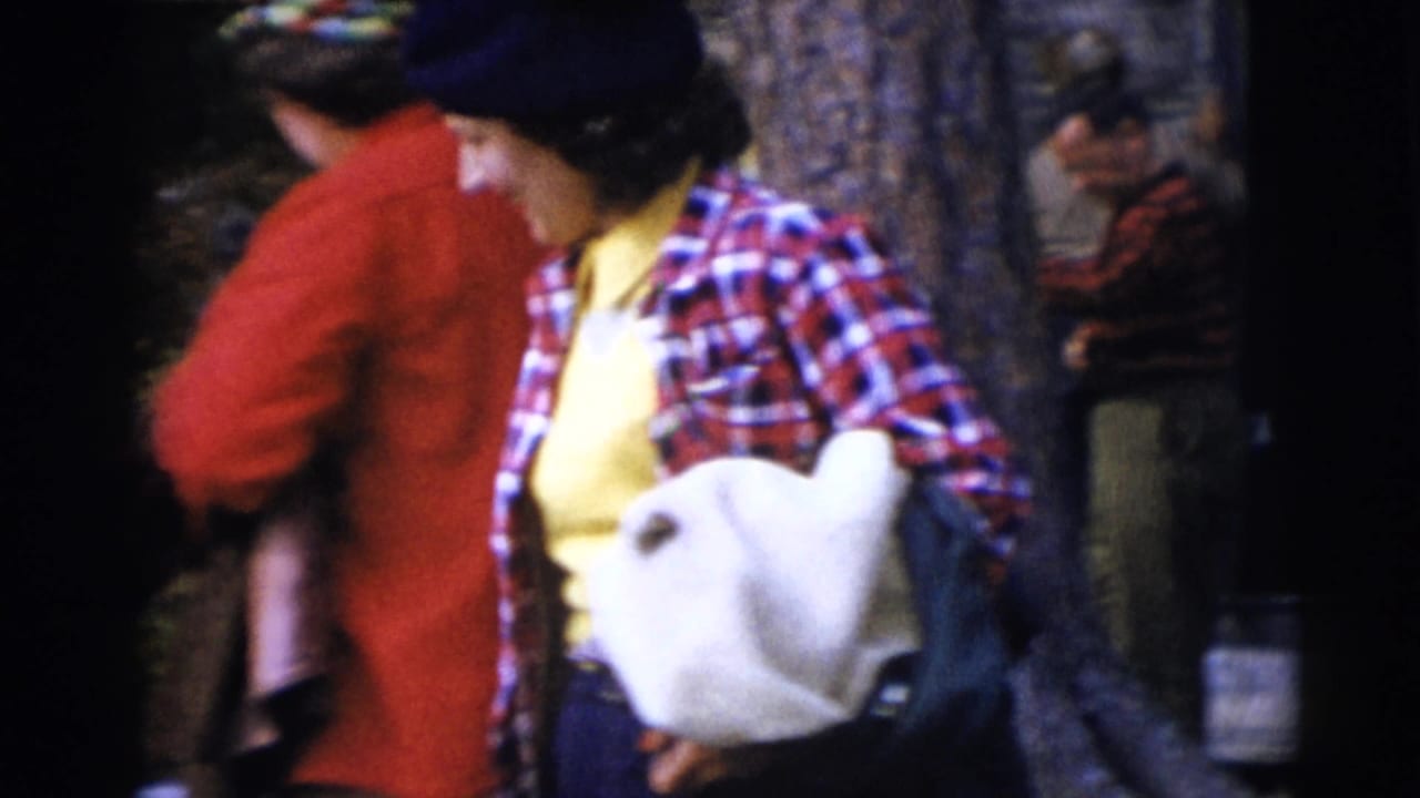 Adults in plaid and outdoor clothing stand near a rustic cabin area in 1953 Baraboo footage, with a woman carrying a white bag or cushion.