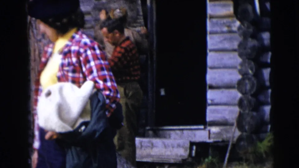 A woman in a plaid shirt and beret carries a white bag beside a log cabin doorway in the Baraboo, Wisconsin home-movie scene.