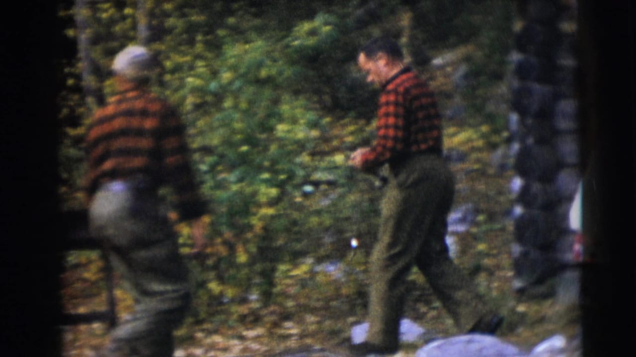 Two men in plaid shirts walking through a wooded area with rocks and leaves.