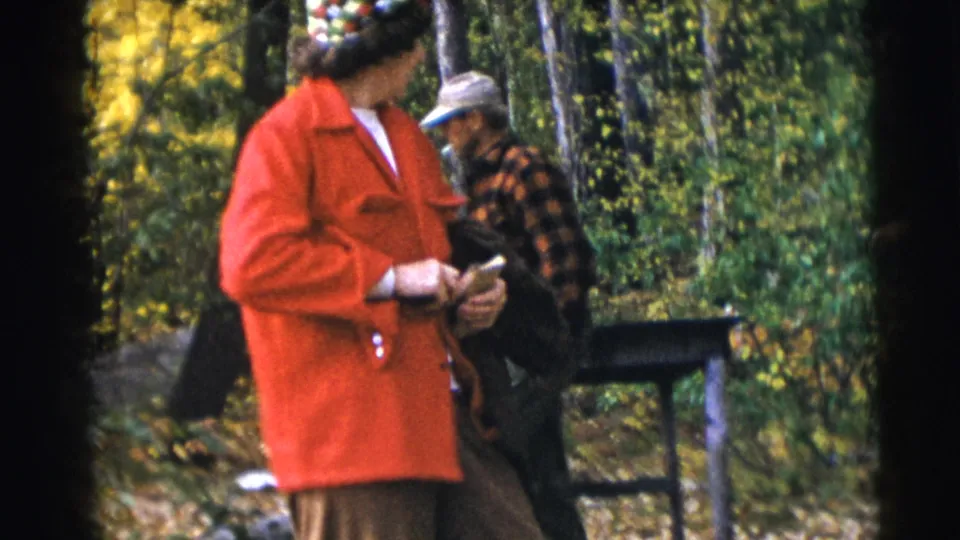 A woman in a bright red coat stands in the wooded Baraboo scene while a man in plaid is partly visible nearby.