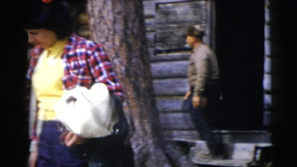 Woman in plaid and a yellow top walks with a white bag near a log cabin in the Baraboo, Wisconsin home-movie scene.