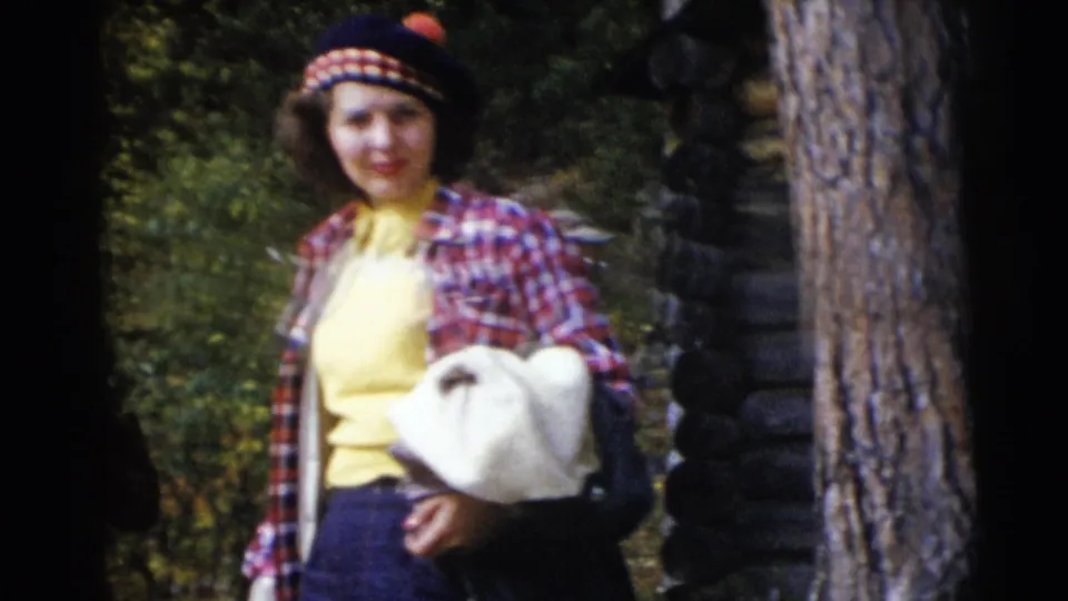 Woman in mid-century casual clothing stands near trees and a log cabin, holding a white bag during the 1953 Baraboo outing.