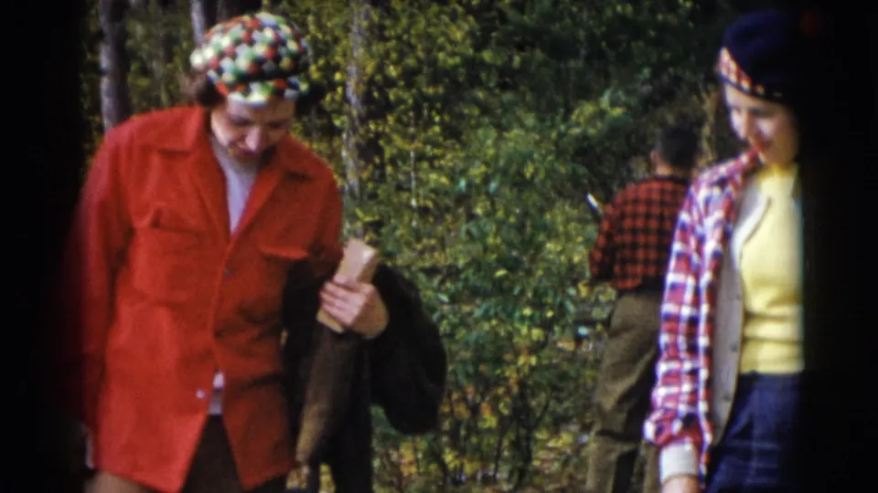 Man in a red jacket and patterned hat carries a rolled item as a woman in plaid passes nearby in wooded Baraboo footage.