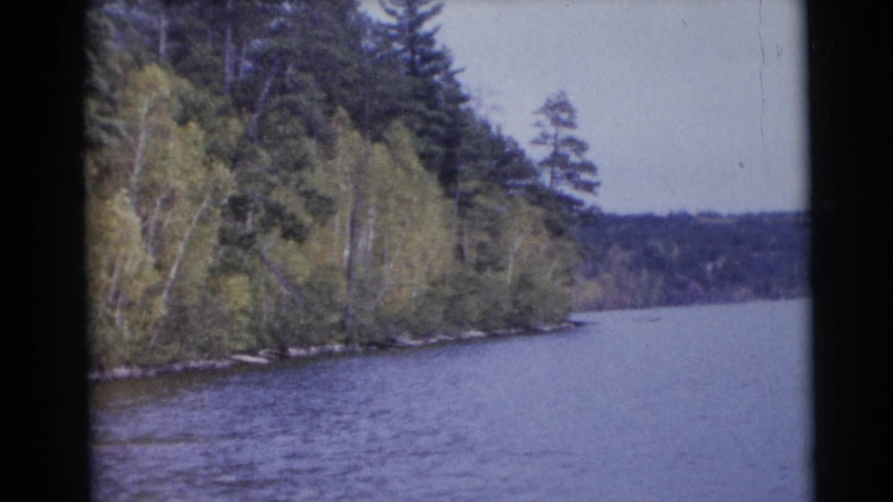 1953 Baraboo, Wisconsin 8mm frame of a river passing a dense forested bank, with distant low hills and a pale sky.