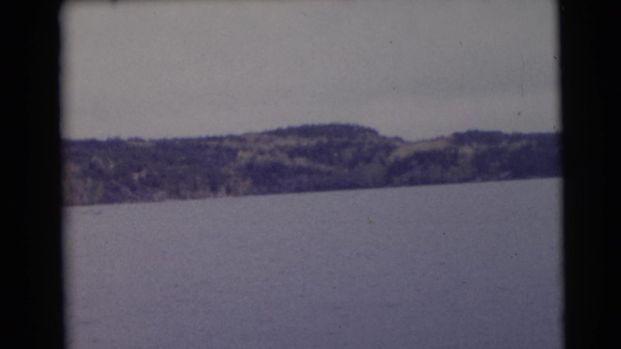 Wide lake surface with distant forested hills beneath a pale overcast sky.