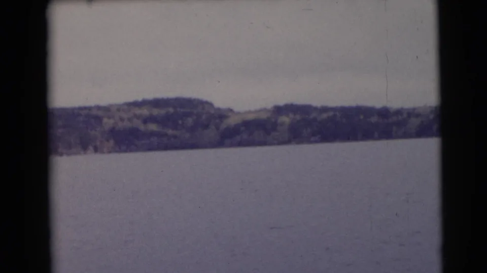1953 Baraboo, Wisconsin 8mm still of lake water below a distant tree-covered hillside and cloudy sky.