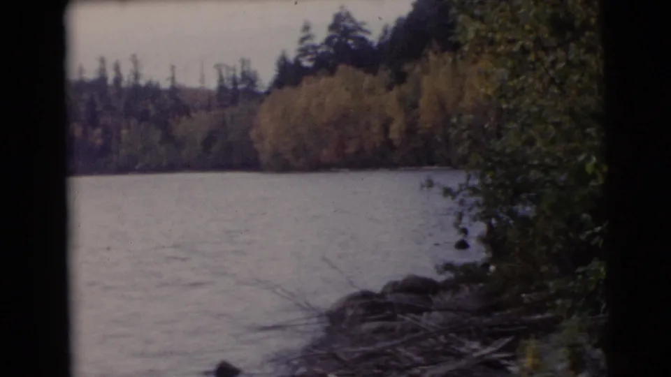 1953 Baraboo, Wisconsin 8mm view of a calm lake bordered by trees, brush, shoreline rocks, and early autumn foliage.
