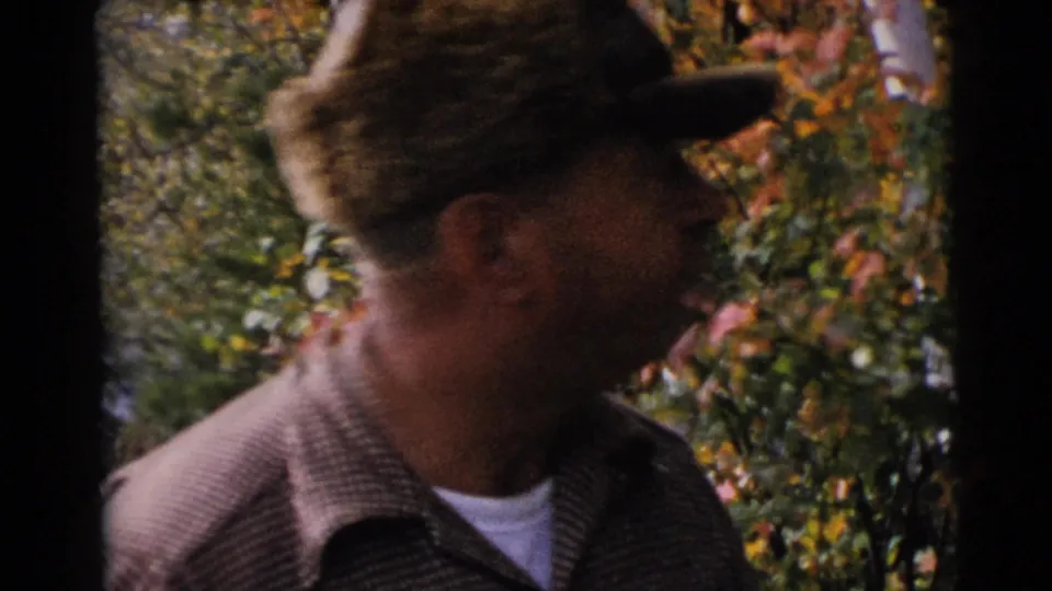 Adult man in profile near the Baraboo lake shore, wearing a fur-trimmed cap and plaid jacket against colorful foliage.