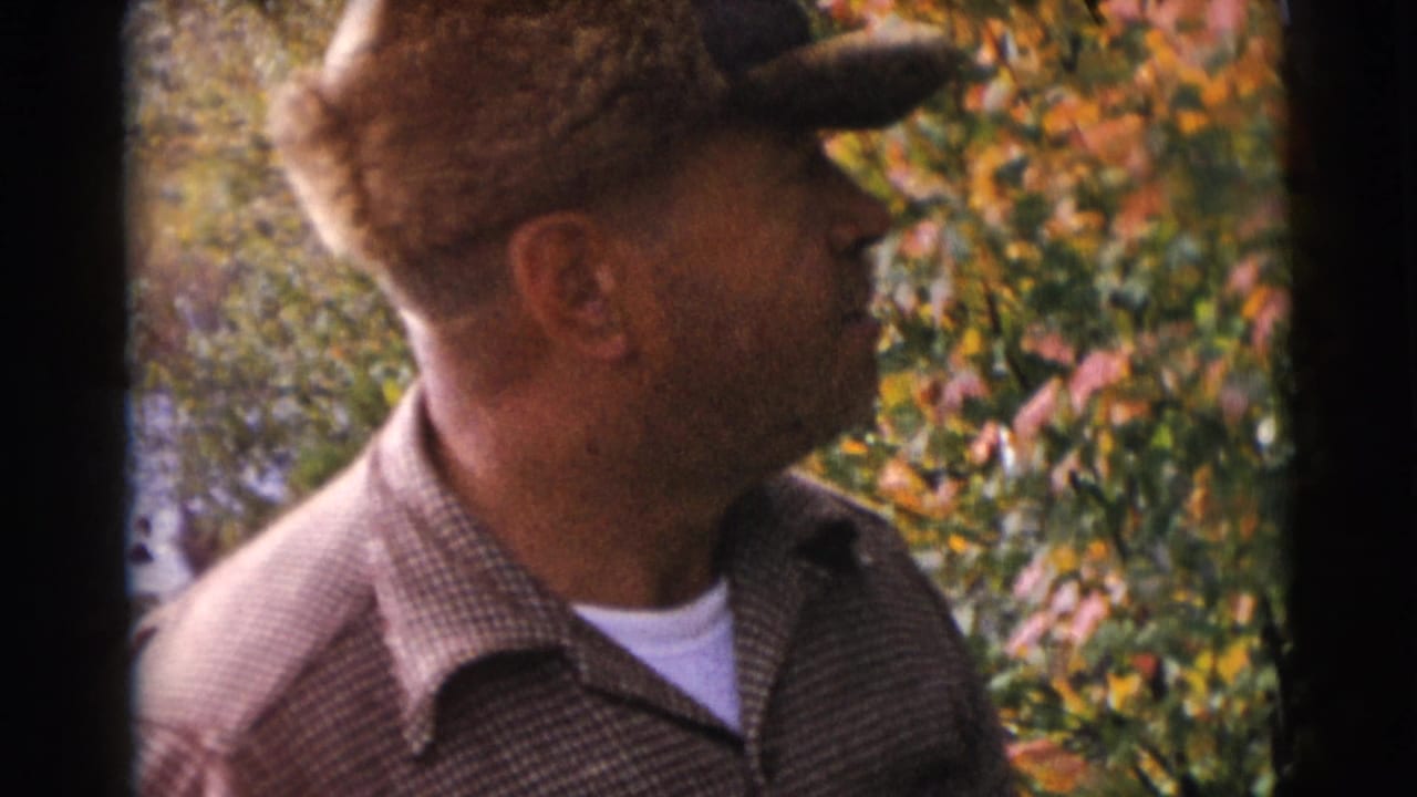 In this 1953 Baraboo, Wisconsin 8mm frame, a man in a fur-trimmed hat and checked jacket stands outdoors beside colorful foliage.