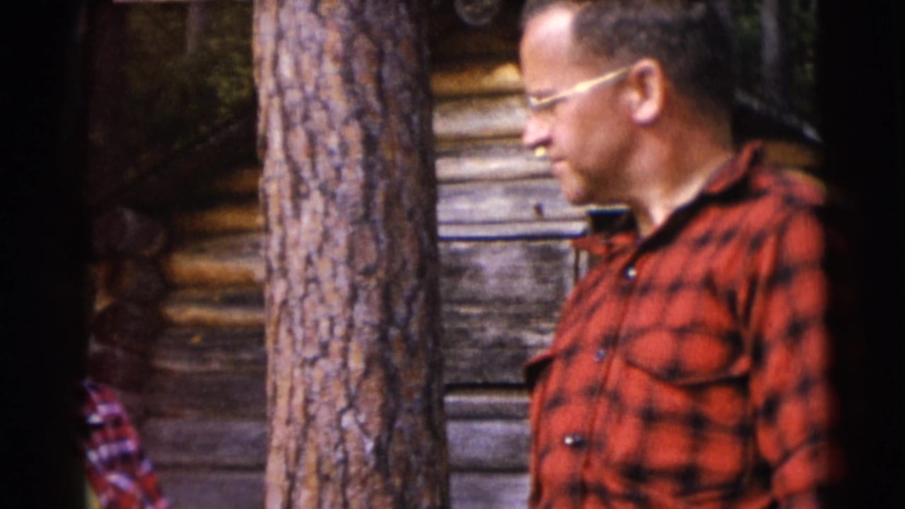 A man in a red flannel shirt and glasses stands outside by a tree trunk and rustic log wall in Baraboo, Wisconsin home-movie footage.