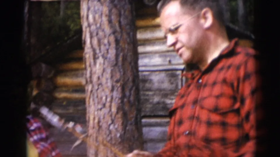 The red-flannel man remains near the tree and log wall, with a partly visible person in plaid at the lower left of the frame.