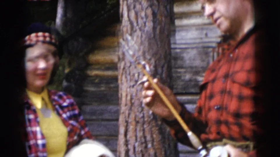 A woman in plaid and a patterned hat stands near the log wall while the red-flannel man holds a long object in the foreground.