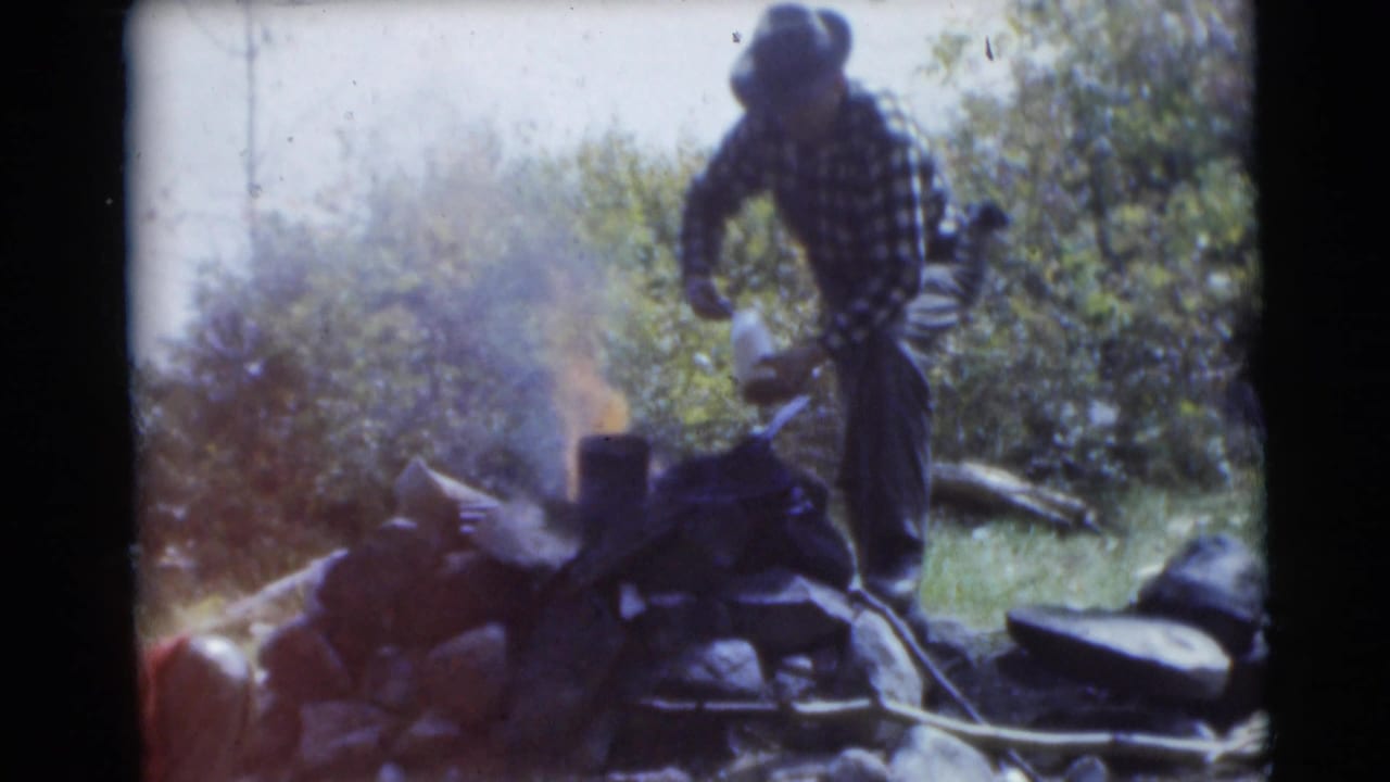 In 1953 Baraboo, Wisconsin footage, a man in plaid flannel and a hat tends a small campfire at a wooded campsite.