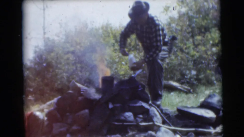 In 1953 Baraboo, Wisconsin footage, a man in plaid flannel and a hat tends a small campfire at a wooded campsite.