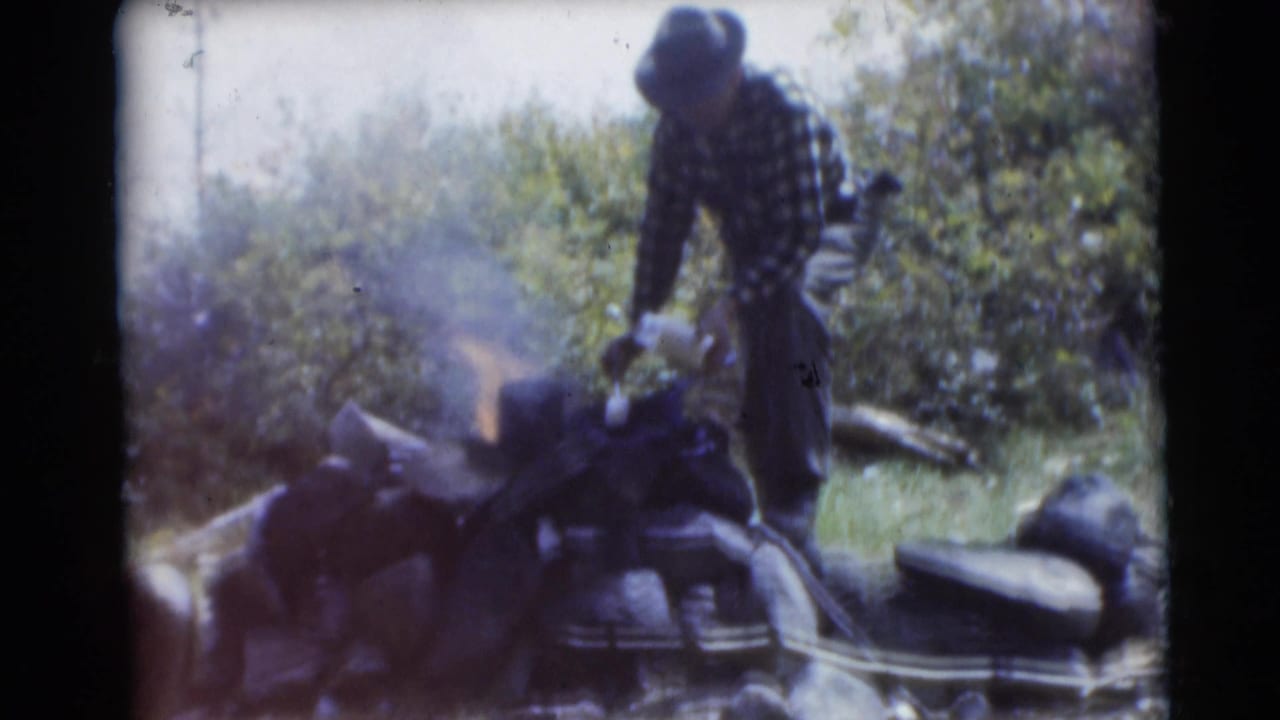 Man stands over a smoking campfire with bags and brush around him.