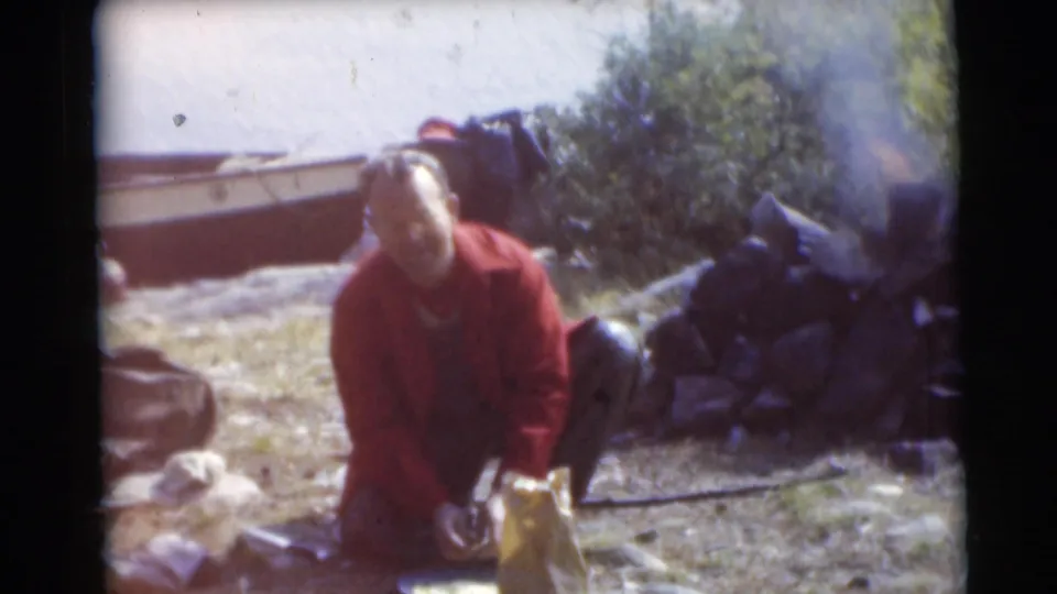A man in a red jacket crouches by supplies at the wooded Baraboo campsite, with a small boat visible behind him.