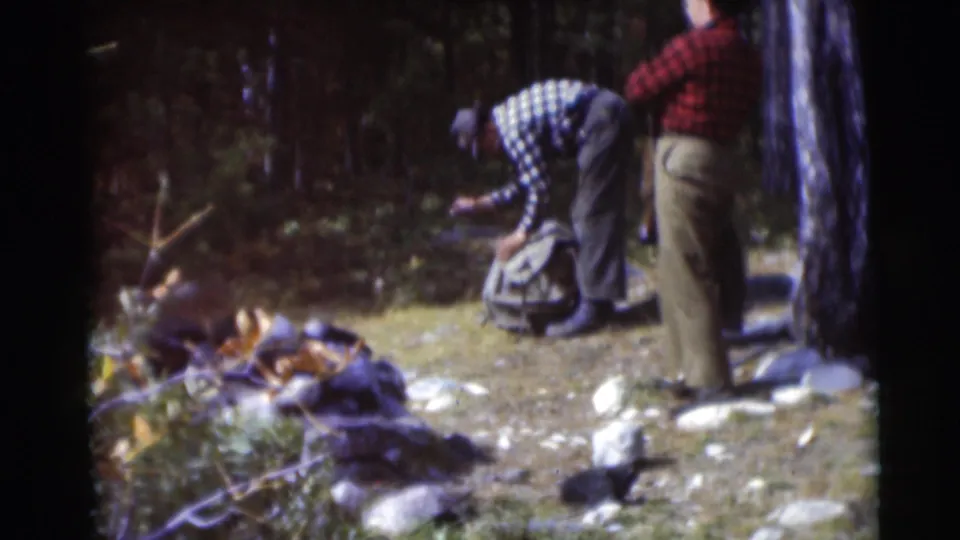 Two adult men in a Baraboo, Wisconsin wooded clearing in 1953, one bending toward a backpack while the other stands nearby.