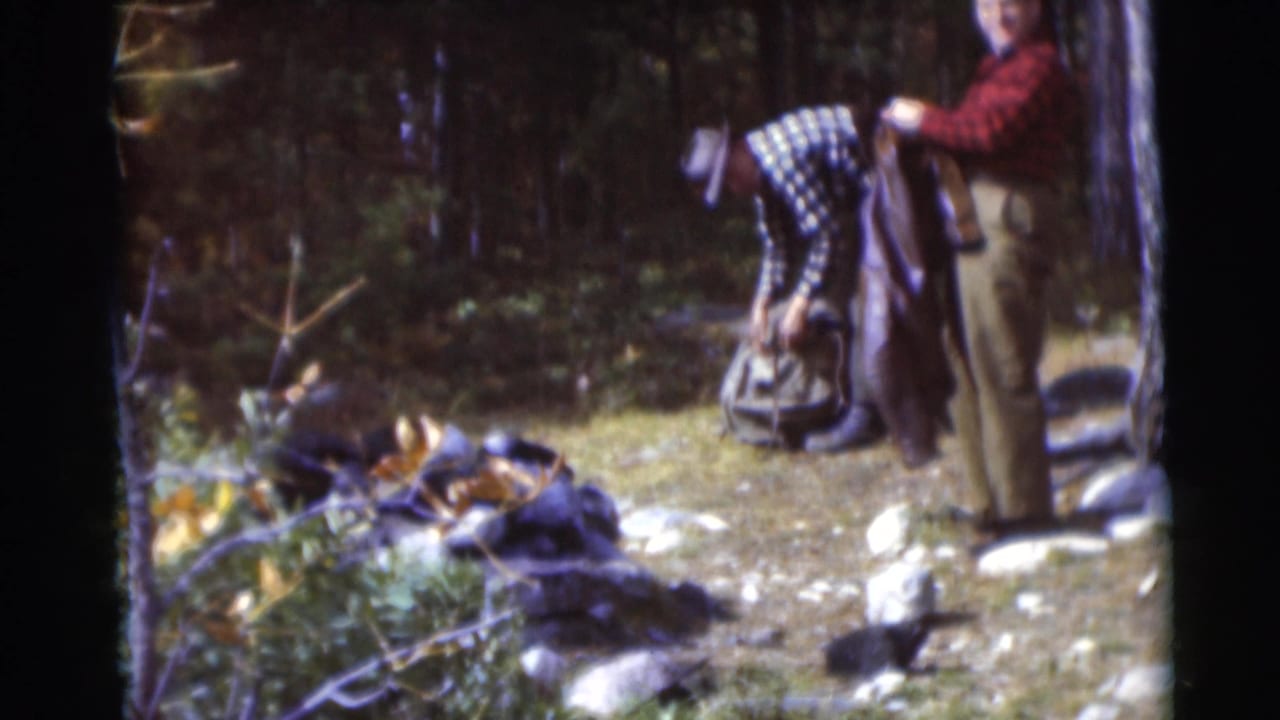 In the 1953 Baraboo woods scene, one man leans toward a backpack as another stands beside a tree holding clothing or gear.