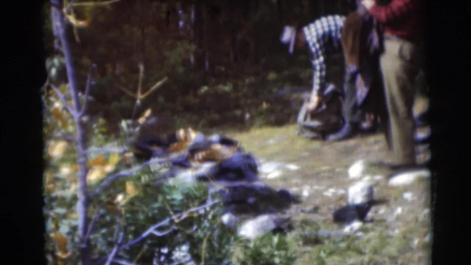 Two adult men in a 1953 Baraboo wooded clearing handle gear near rocks and brush, with the framing partly cropped by the 8mm scan.