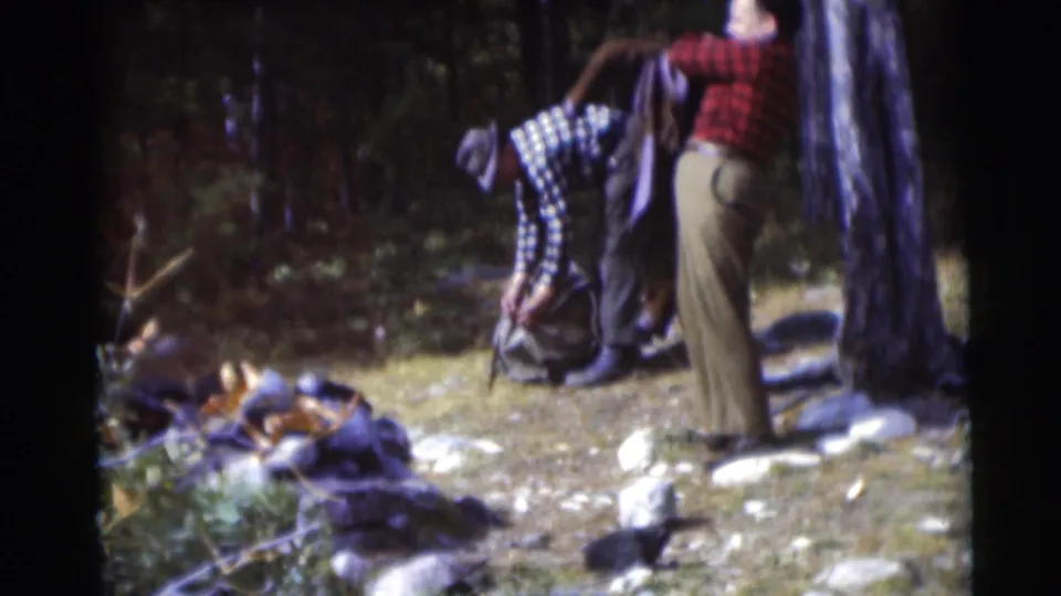 A man in a red plaid shirt stands near a tree while another man bends over a backpack in the 1953 Baraboo wooded setting.