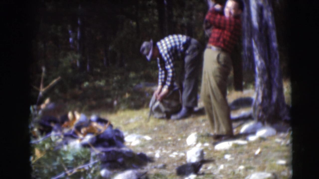 Two older men in plaid shirts stand in a wooded clearing as one bends over a canvas bag.