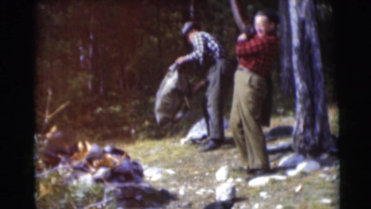 A man lifts or packs a large canvas bag while another man stands beside a tree.