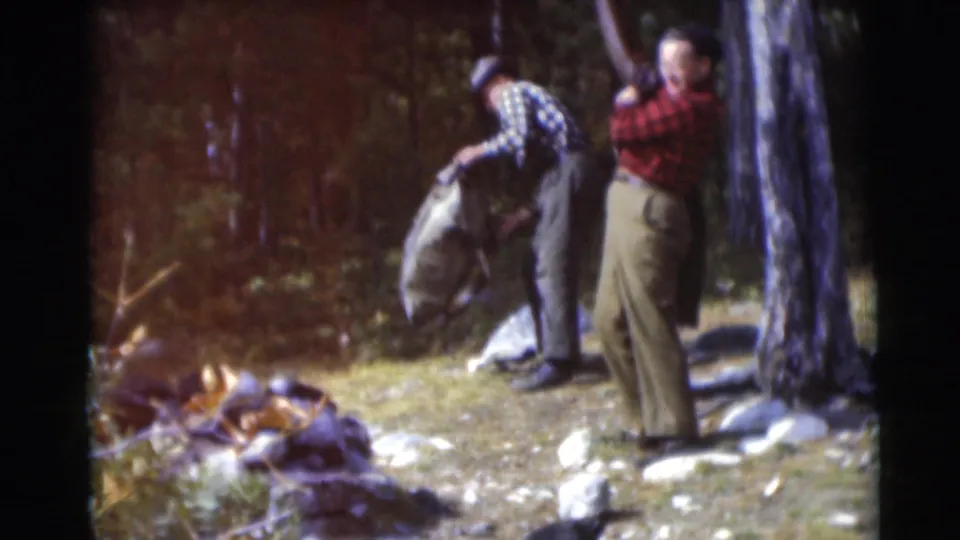 Two older men in a 1953 Wisconsin forest clearing handle outdoor gear, with one lifting a large canvas bag near a tree.