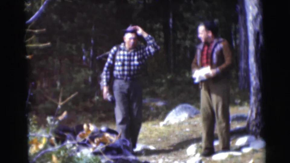 In the wooded Baraboo setting, one older man adjusts his hat while another holds a paper or map in 1953 home-movie footage.