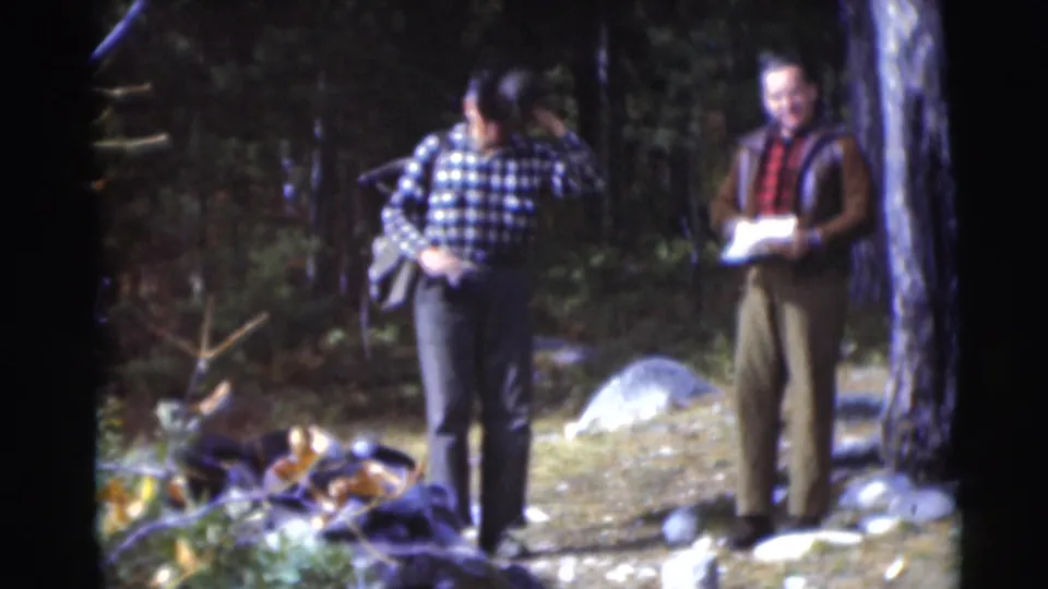 Two older men in plaid clothing pause in a 1953 Baraboo forest clearing, with a bag and paper visible as outdoor gear.