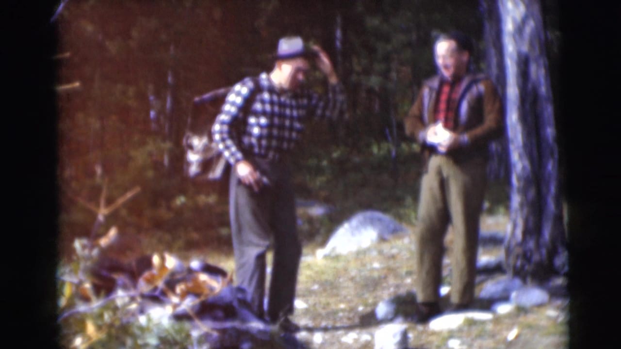 Two men stand in a wooded area, one adjusting his hat while wearing a backpack.