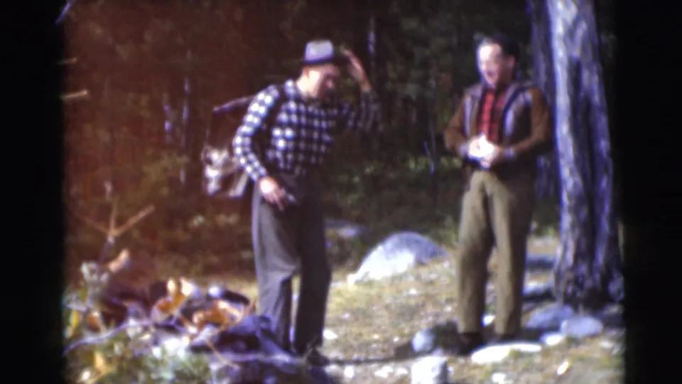 In 1953 Baraboo, Wisconsin footage, two adult men stand among trees and rocks as one adjusts his hat and carries a backpack.