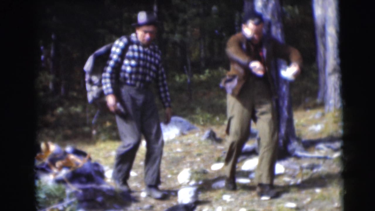 In the wooded Baraboo scene, a man in a checkered shirt and backpack stands near another man handling a white object.