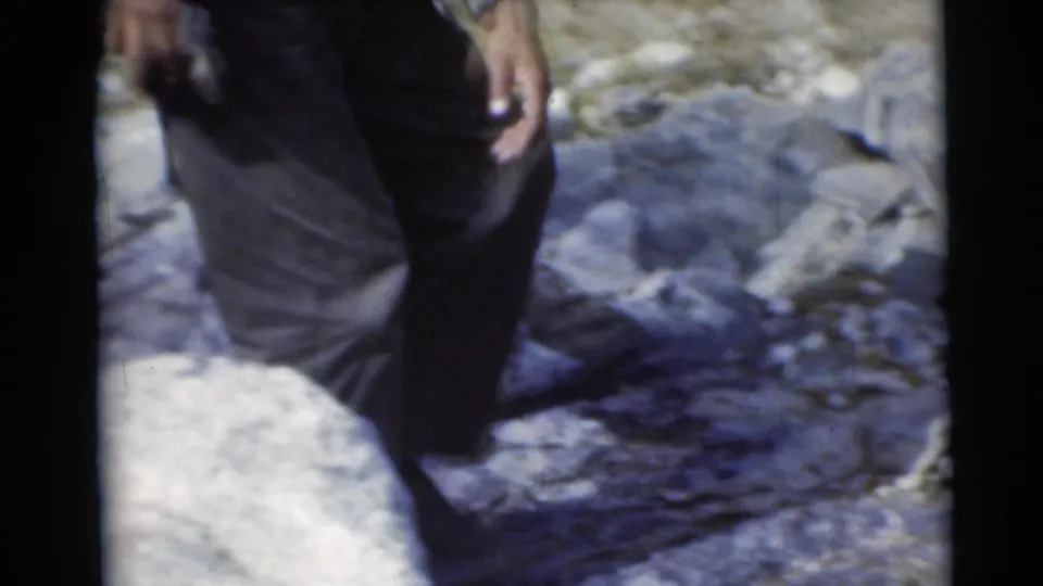 A man’s legs and hand pass through a rocky woodland area in the 1953 Baraboo camping home movie.