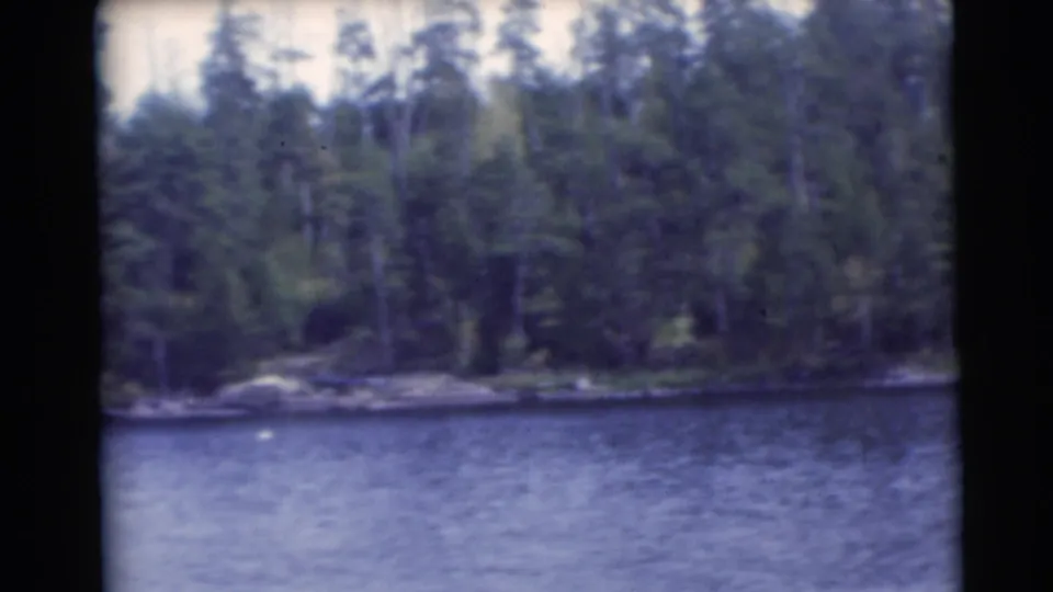 1953 Baraboo, Wisconsin 8mm frame with mixed green forest, a narrow shore, and blue water in the foreground.