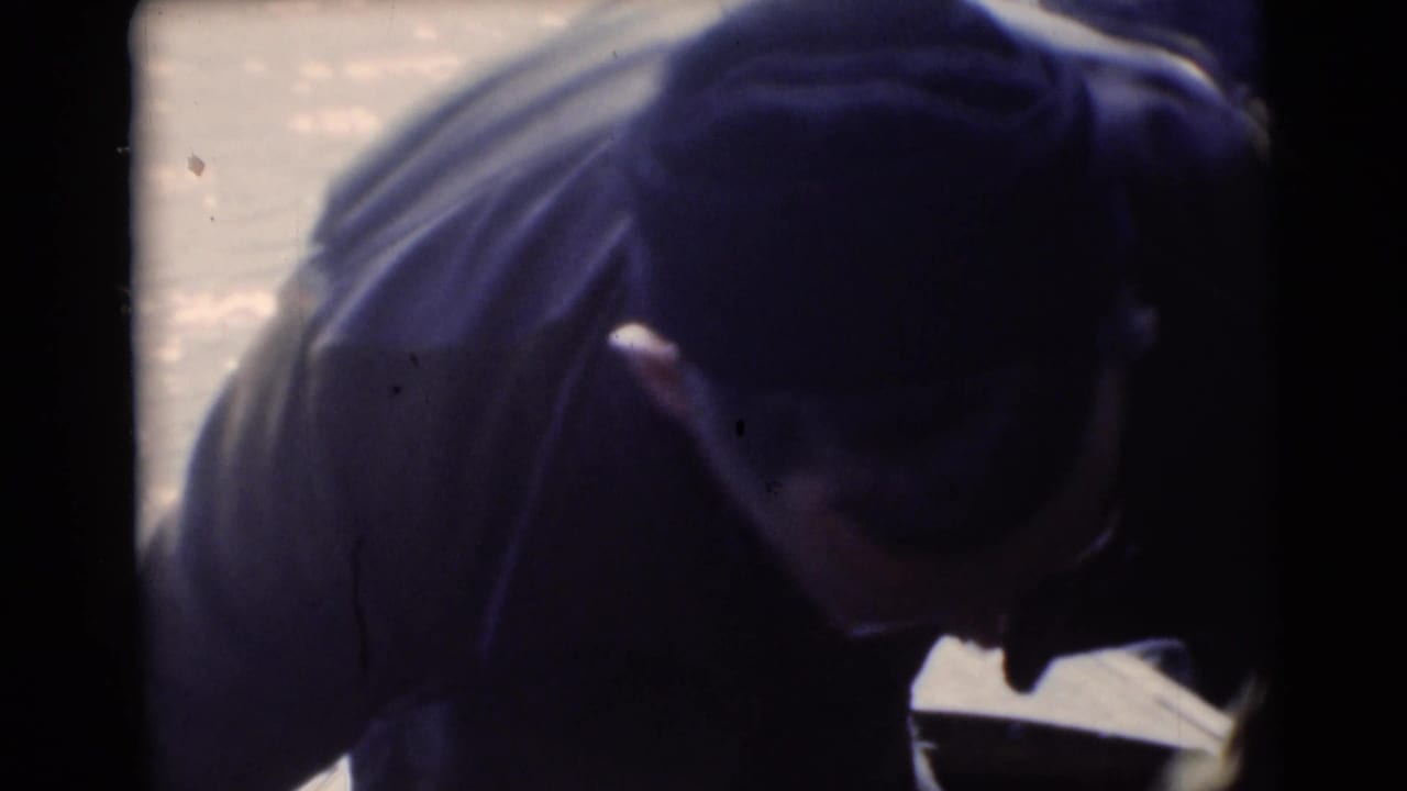 Man in cap and dark jacket bends near water and canoe gear.