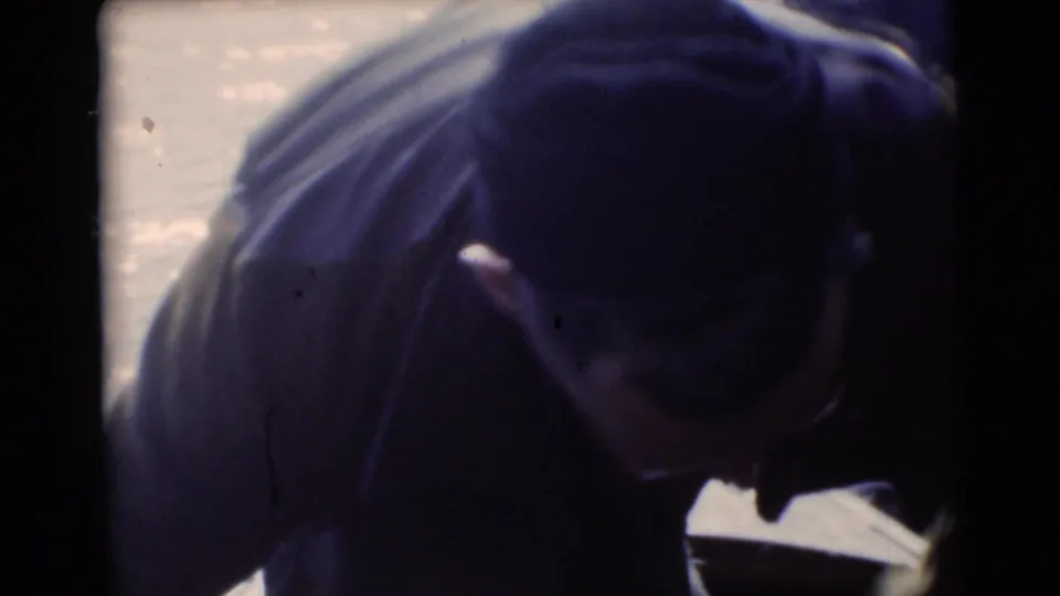 Man in a cap and dark jacket bends near canoe equipment beside the river in 1953 Baraboo home-movie footage.