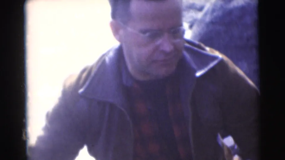 Adult man in a dark jacket stands near the bright riverside area, with his face partly visible in the home-movie frame.