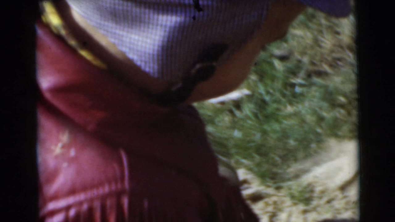 Small child seen from behind in a red fringed jacket and striped cap on grass.