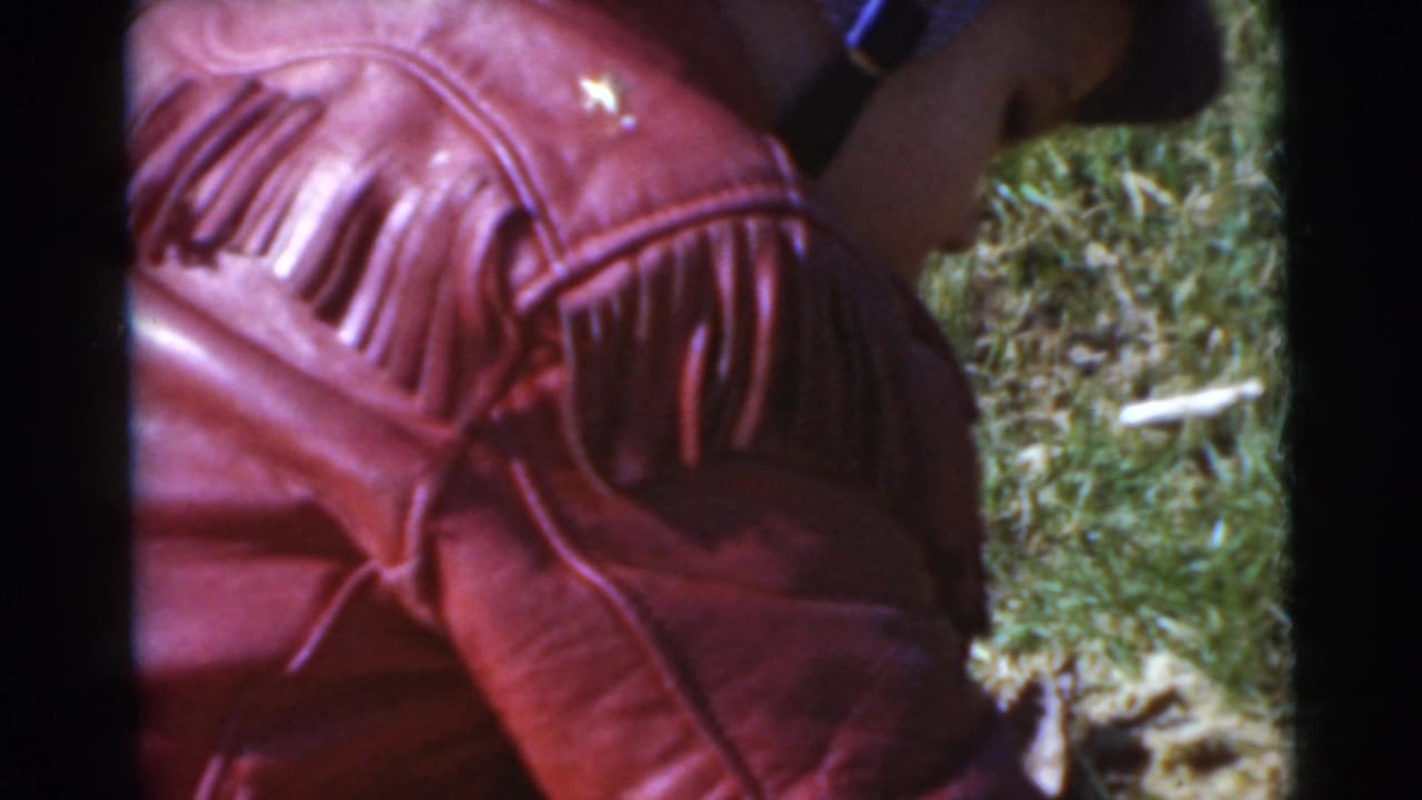 Close 8mm view of the child's red fringed jacket from behind, with grass and a small arm visible in the Miami lawn setting.