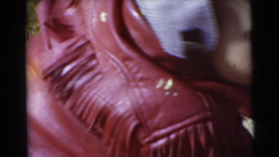 A close, soft 8mm frame shows the child's red fringed jacket, striped cap, and part of an arm against grass in the 1956 Miami footage.