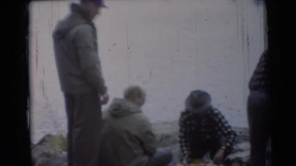 1956 Miami lakeside home-movie frame showing men near the water, including a standing figure and seated figures in casual clothing.