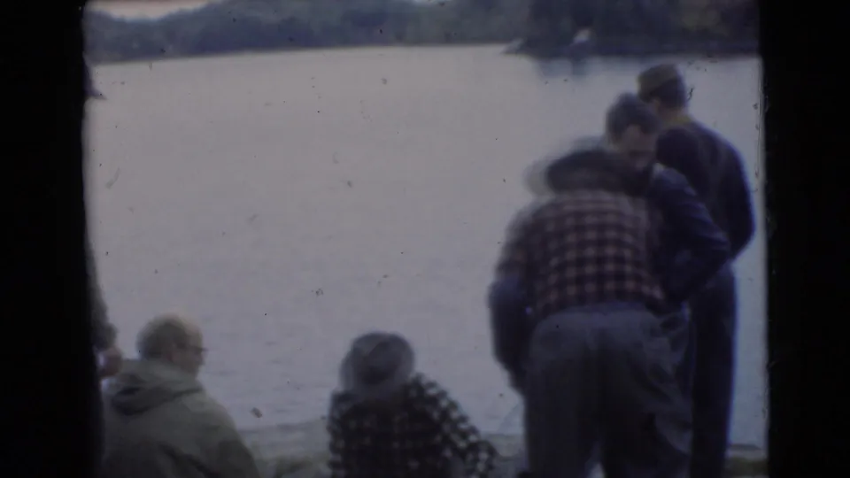 1956 Miami 8mm frame showing men in casual jackets and plaid shirts gathered close to the lakeside, with water behind them.