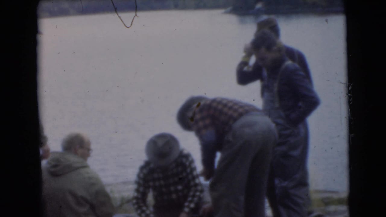 Several men in coats gather by a lake, with one person bending near the shore.