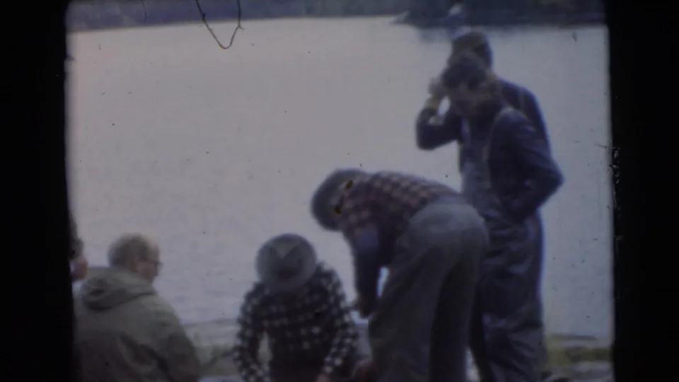 1956 Miami home-movie frame showing adult men in coats and caps gathered beside a lake, some seated and one bending near the water.