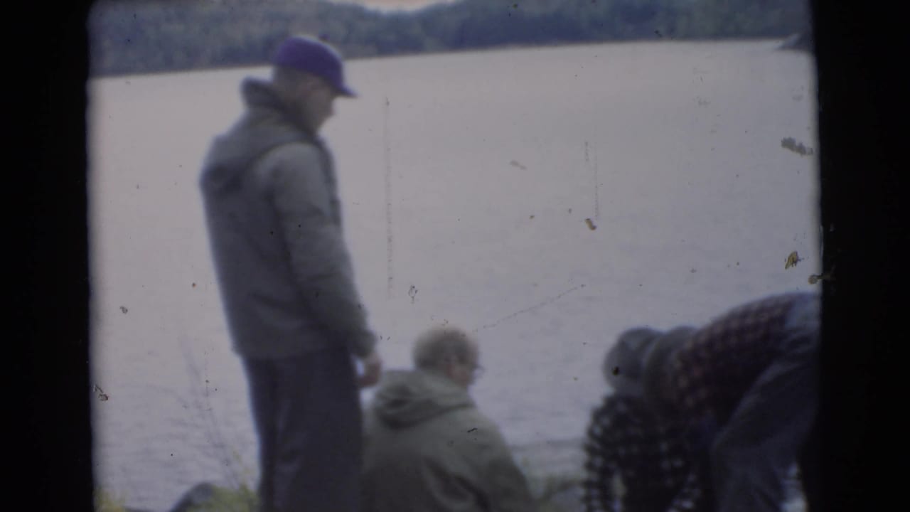 A man in a cap stands near seated men beside the lake.